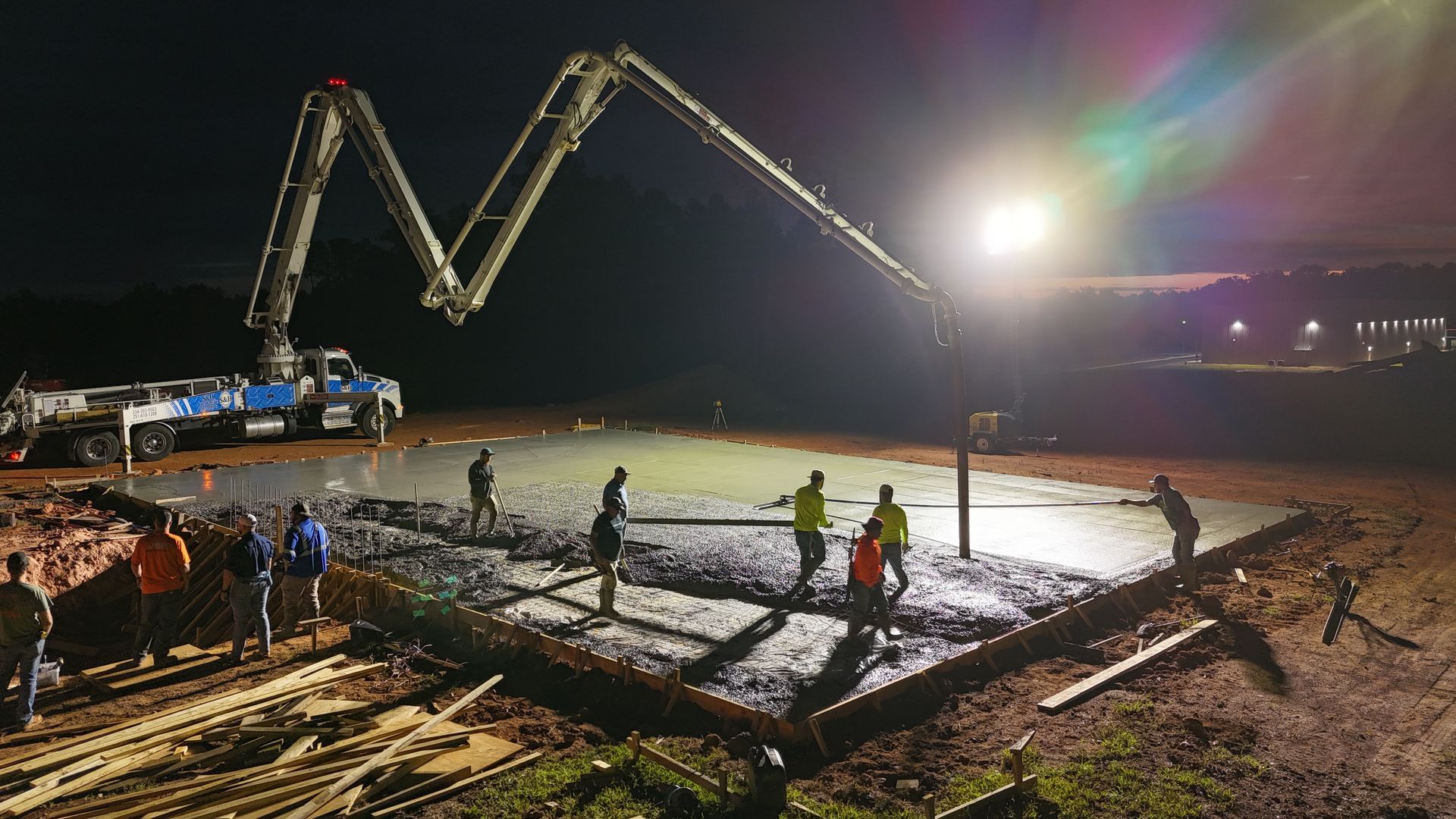 Construction crew pouring concrete at night with a boom pump, floodlights, and workers smoothing a large slab.