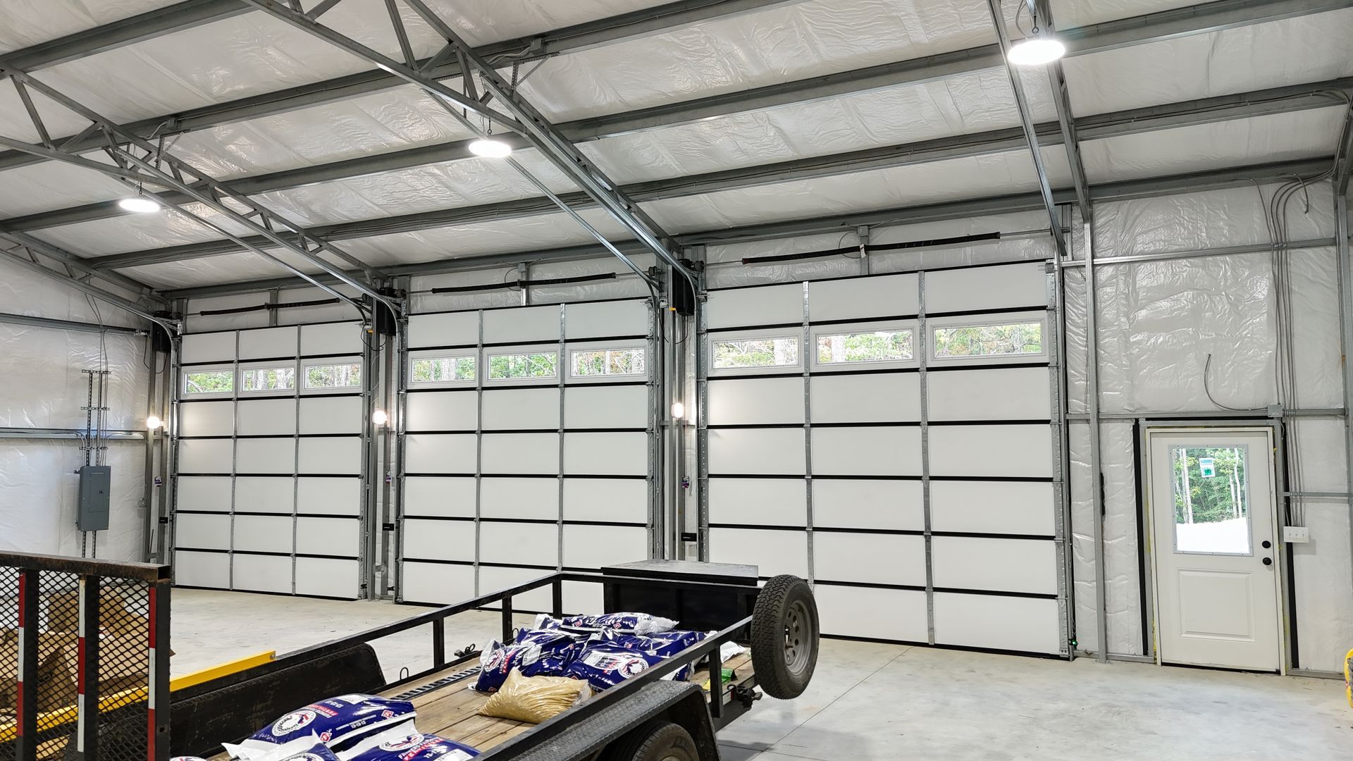 Inside of a large garage with three white doors, a trailer, and a small white door to the right.