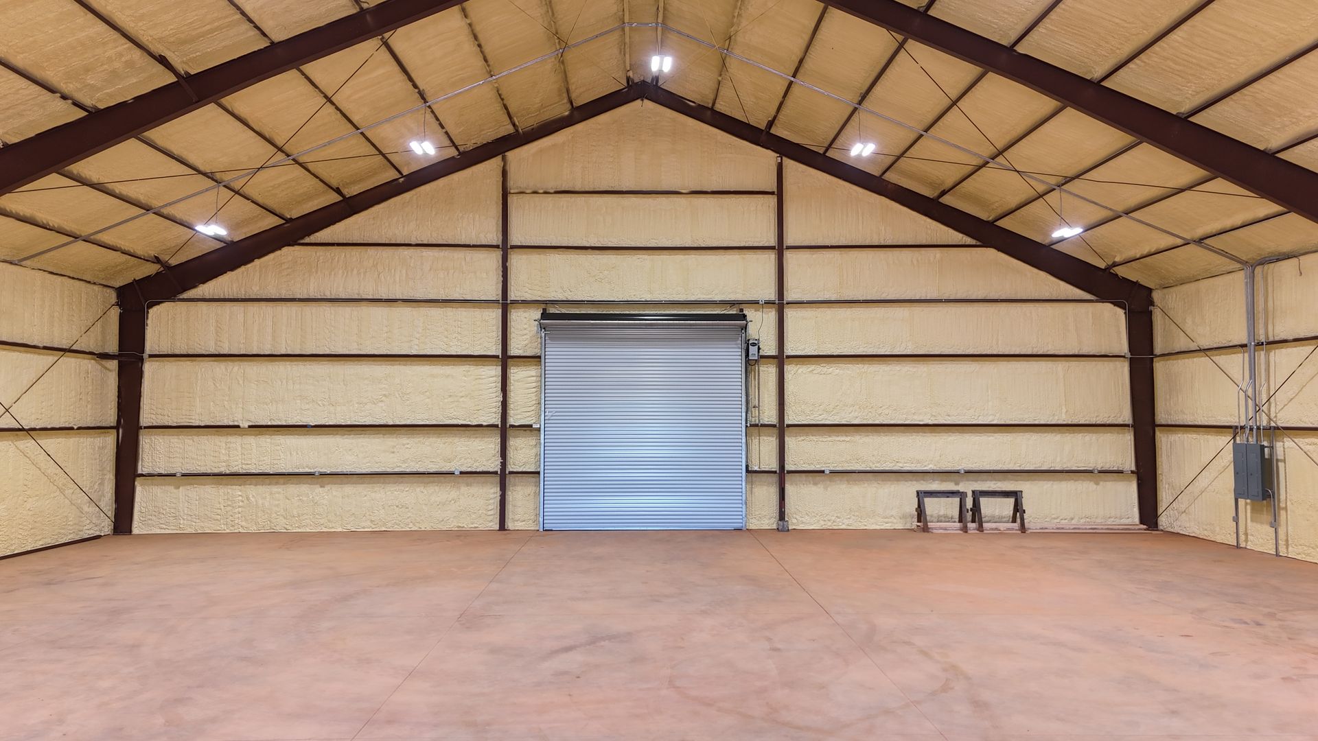 Inside of a tan, open warehouse with a closed garage door. Brown beams and spray foam insulation are visible.