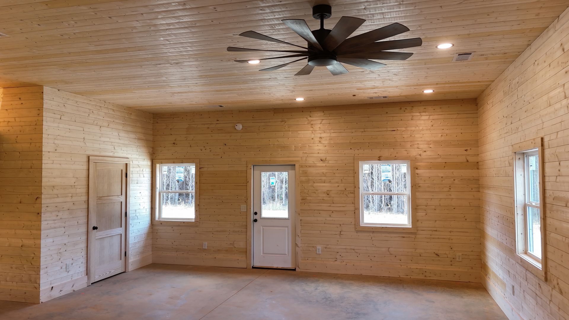 Interior of a room with light wood walls and ceiling, a ceiling fan, and windows.