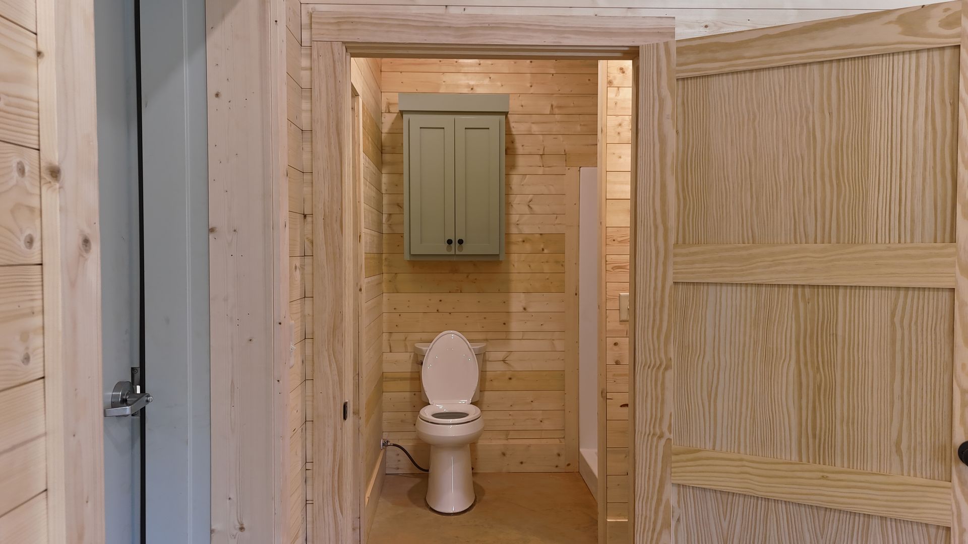 Open wooden door revealing a small bathroom with a toilet, cabinet, and shower. Wood paneling in light tones.