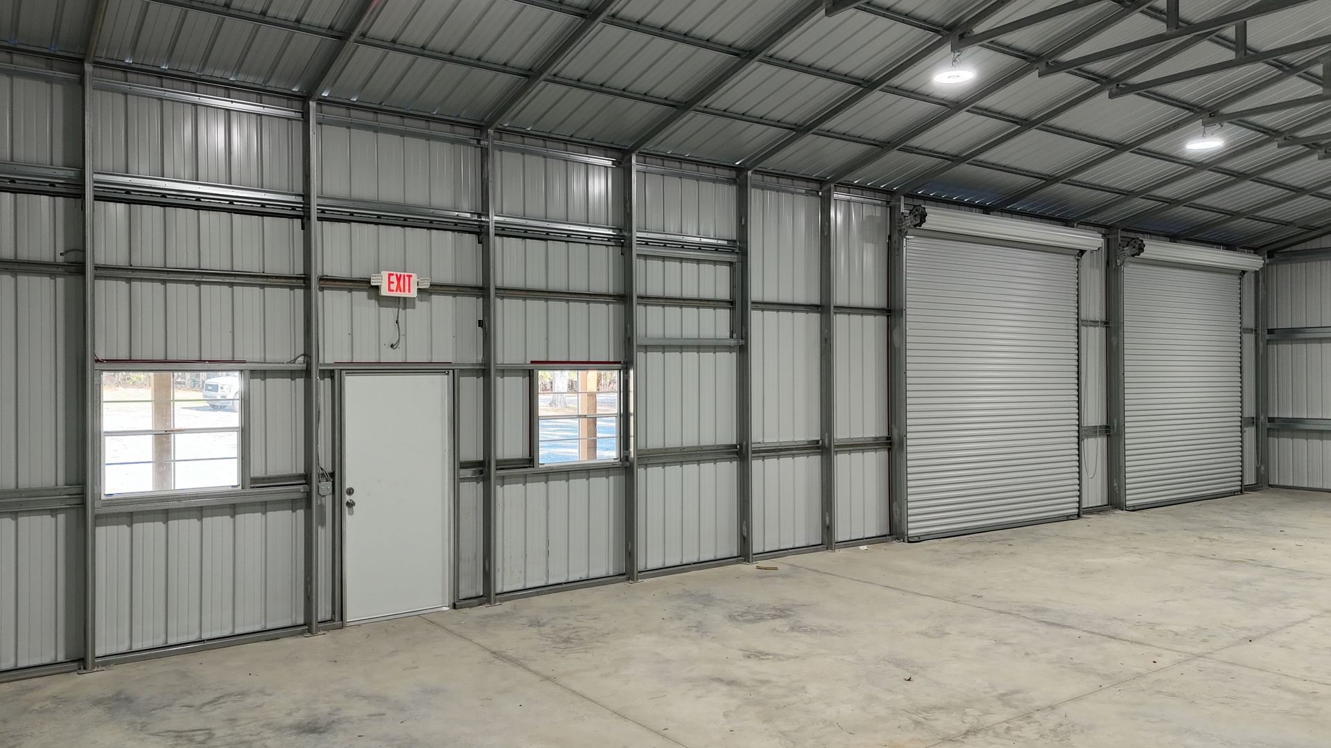 Interior of a large metal warehouse with a roll-up door, exit sign, and windows.