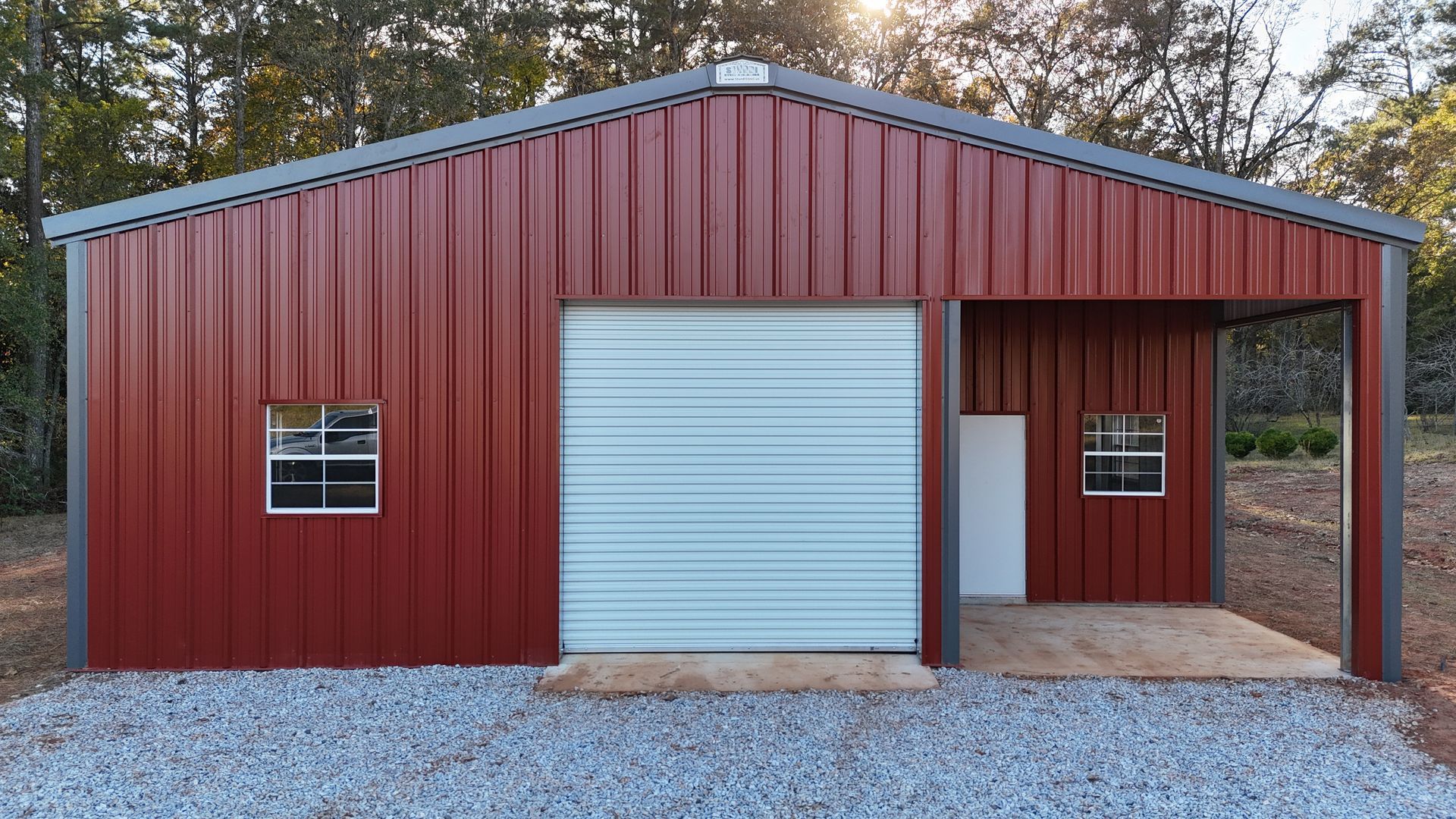 Red metal building with garage door and carport, two windows, gravel ground.