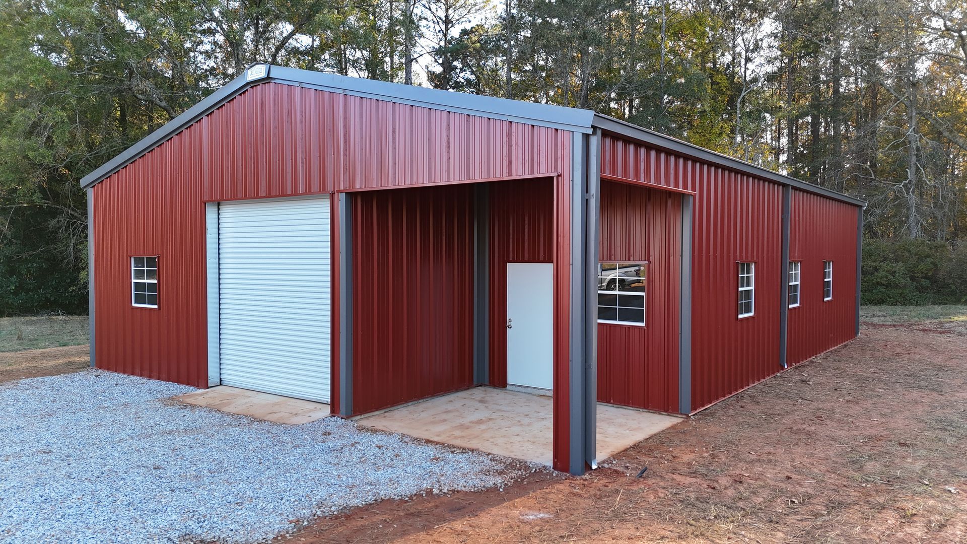 Red metal workshop with garage door, small windows, and open carport. Gravel base, trees in background.
