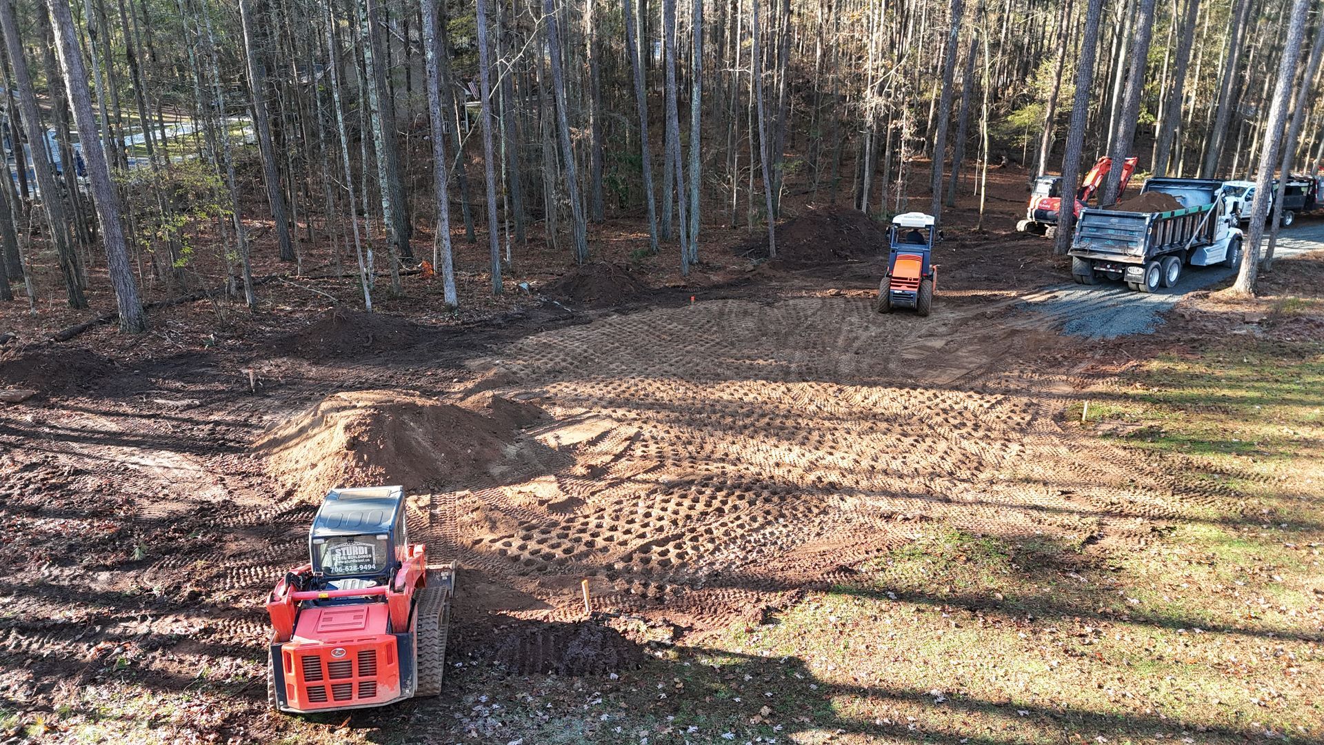 Small tractors working in a wooded clearing with leaf-covered ground and a blue utility vehicle nearby