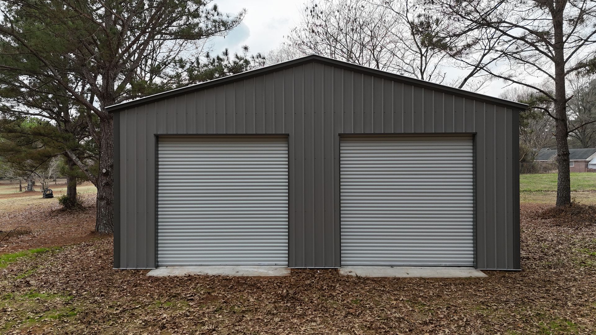 Gray metal garage with two roll-up doors, set in a grassy yard, under a cloudy sky.