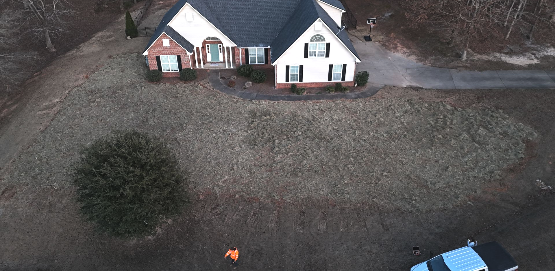 Aerial view of a white house with a dark roof on a large yard bordered by trees.