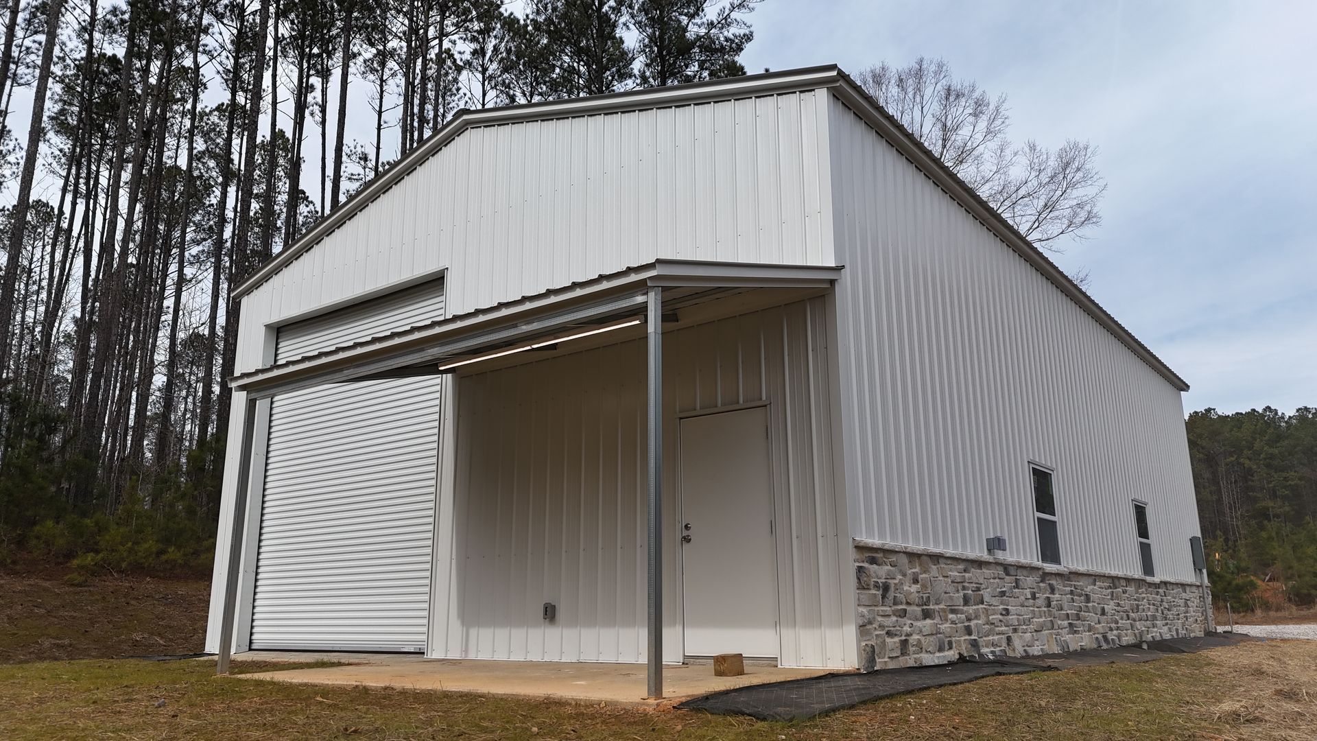 White metal building with open garage door, awning, and stone base in a wooded area.