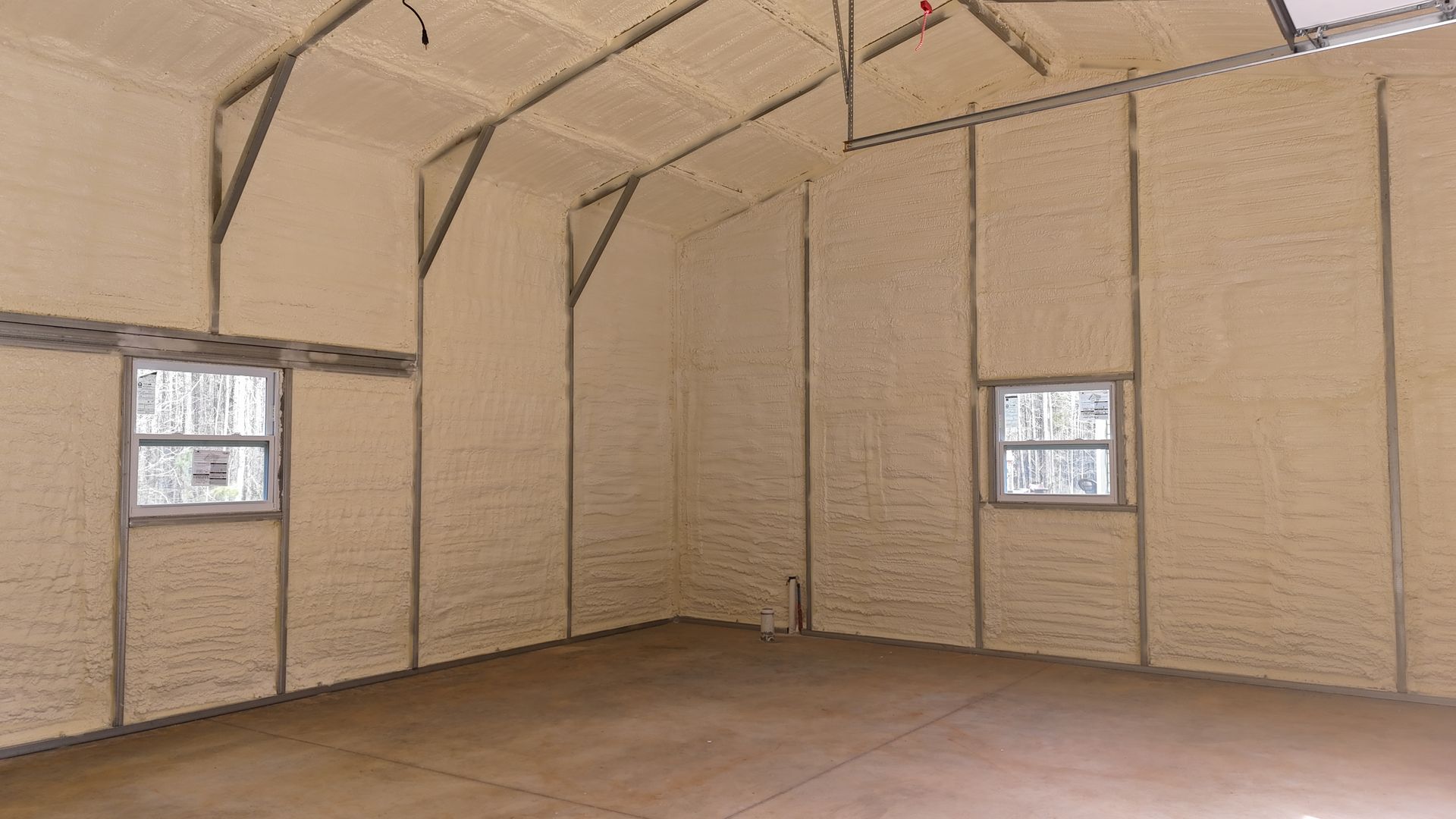 Interior of a building with light tan spray foam insulation on walls and ceiling; two windows, concrete floor.