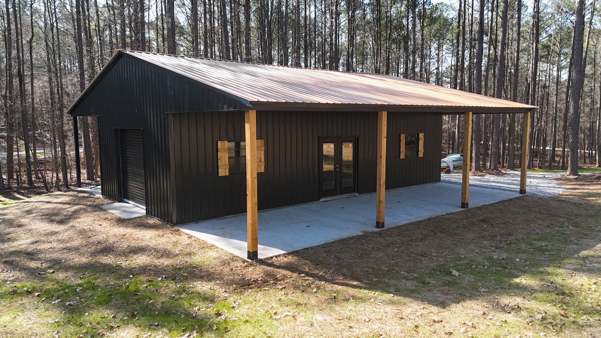 Black building with a corrugated metal roof and covered porch supported by wooden poles, in a wooded area.