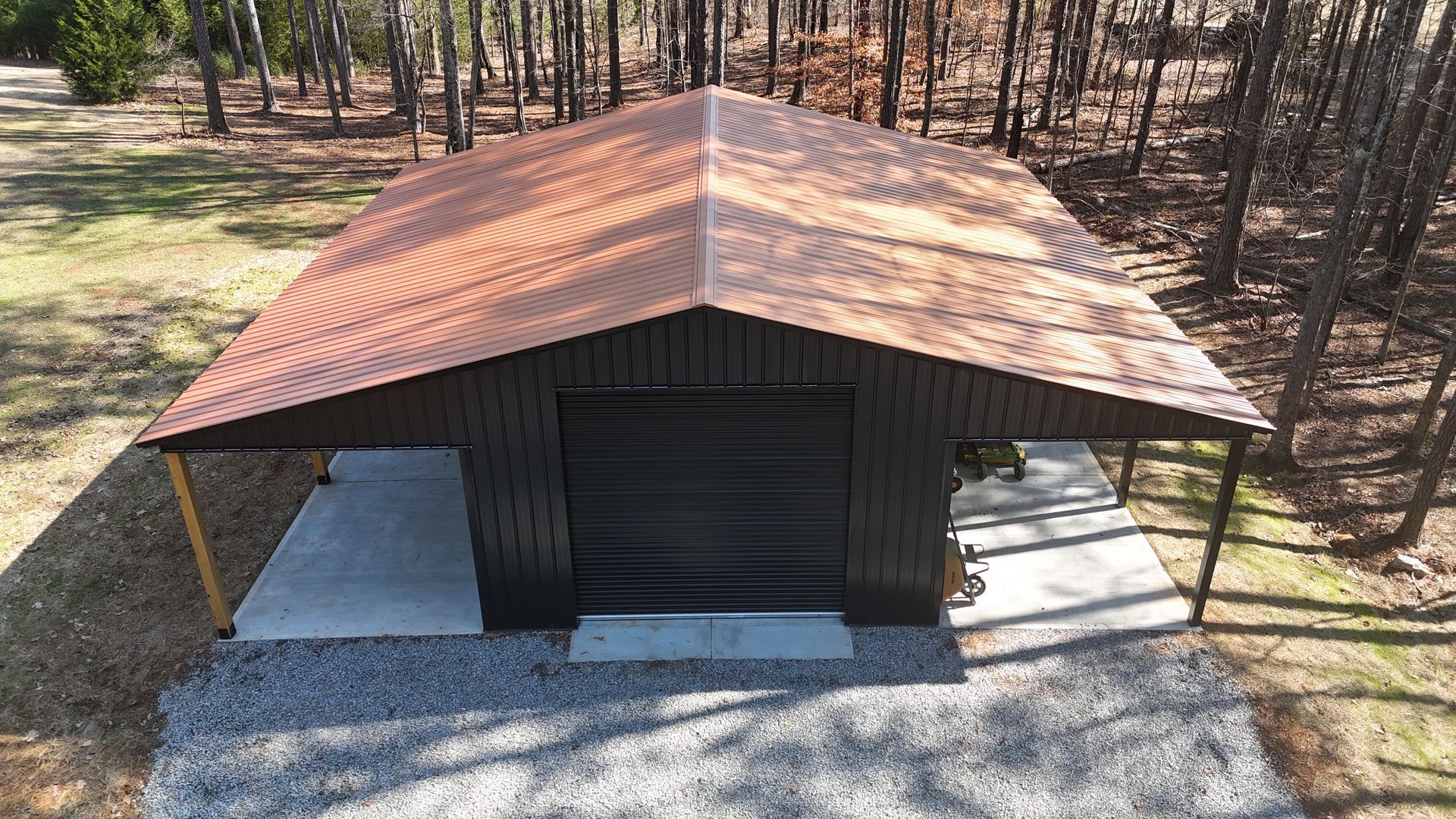 Garage with a copper roof and black siding, on a gravel base in a wooded area.