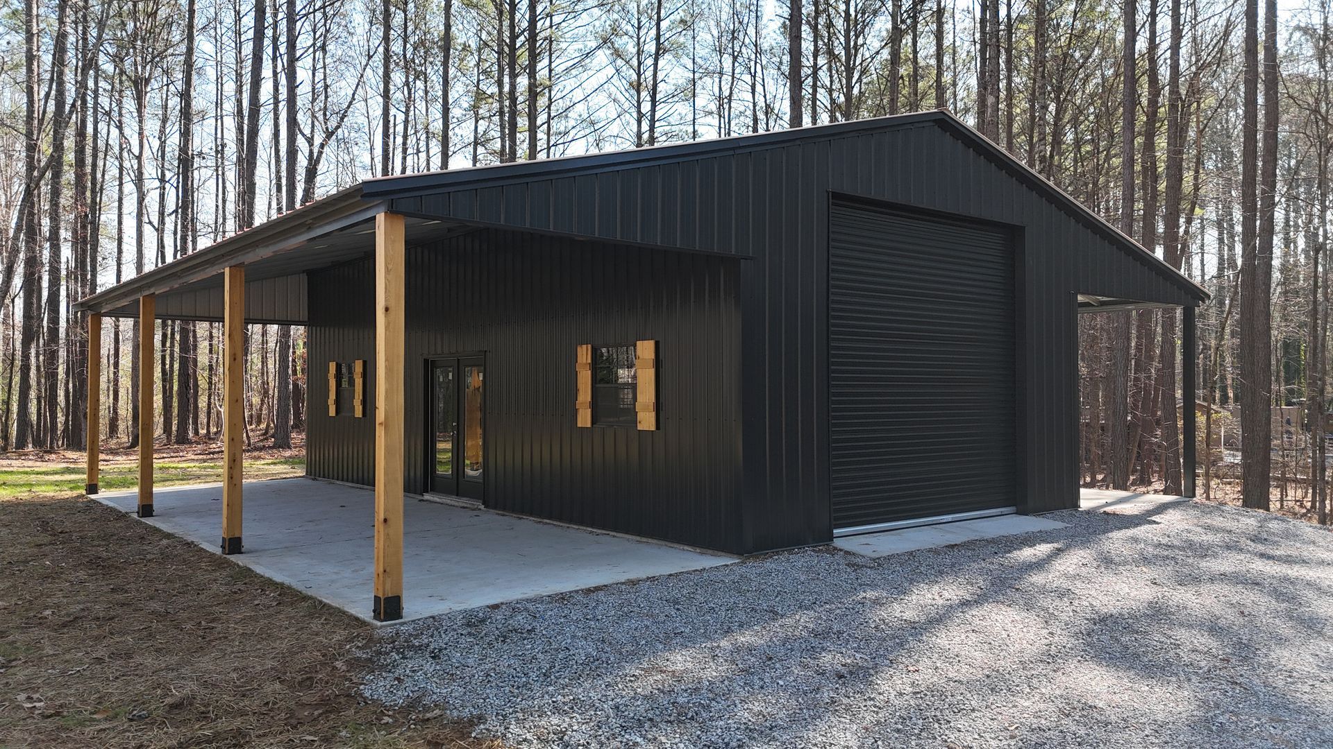Black metal shed with a covered porch and gravel driveway in a wooded area.