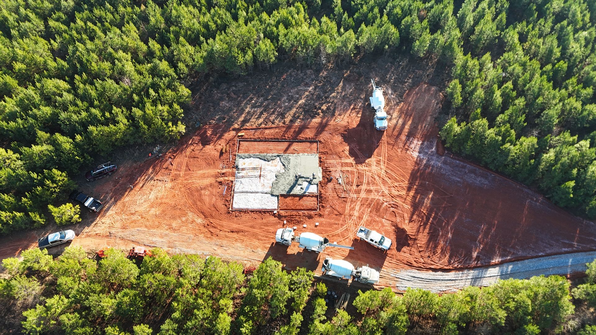 Aerial view of a small construction site with trucks on red dirt surrounded by dense green forest.