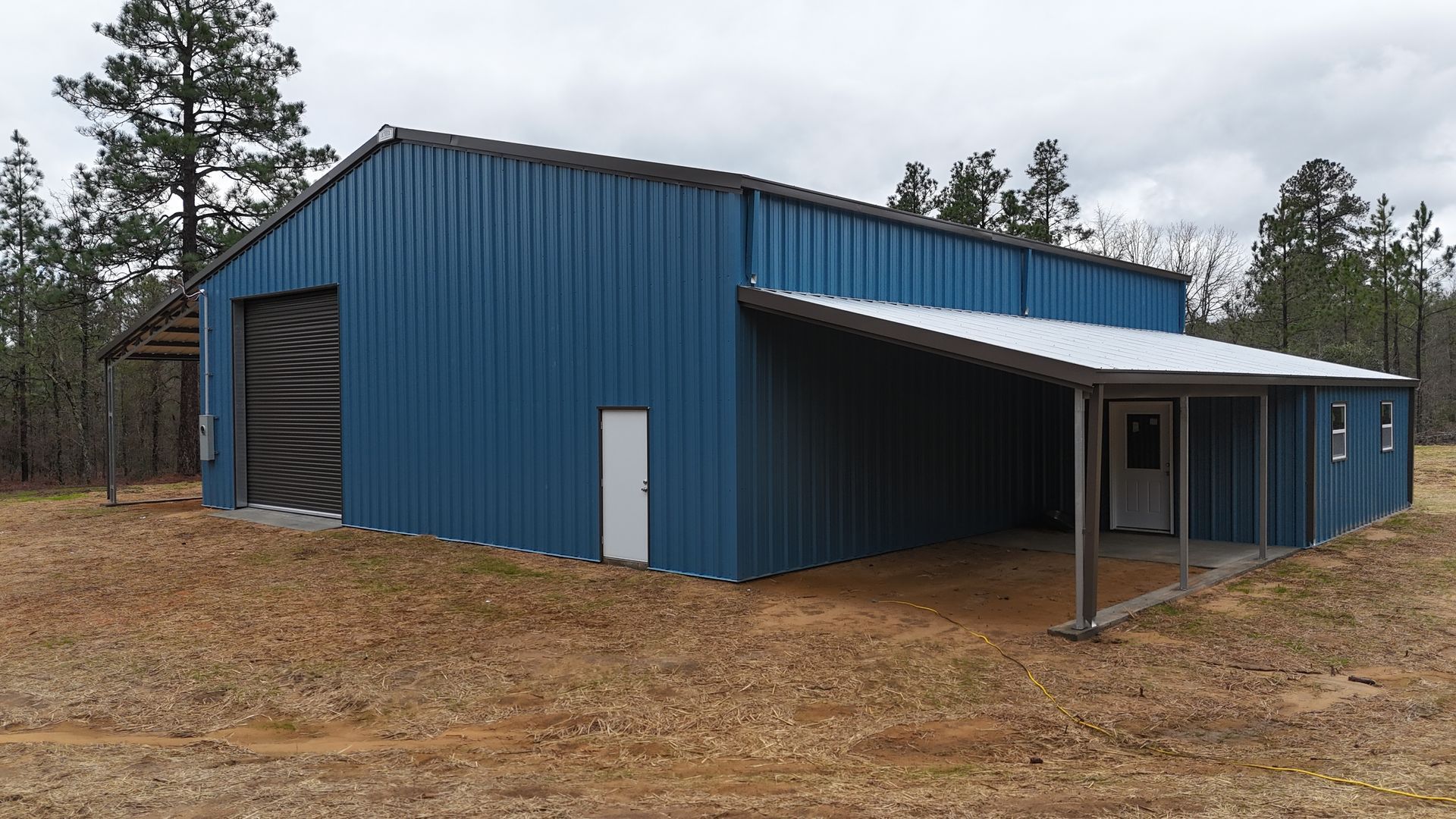 A blue metal pole barn with a roll-up door, side entrance, and an open lean-to porch in a wooded, grassy landscape.