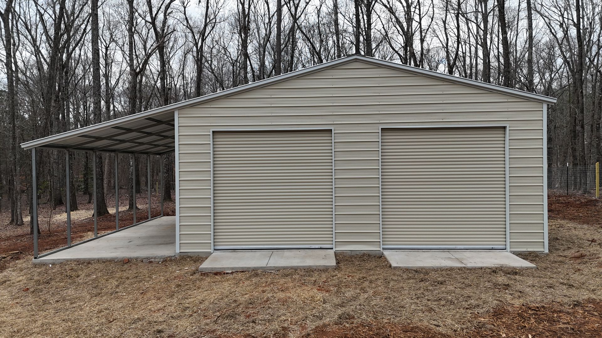 Tan metal storage shed with two roll-up doors and a covered side bay in a wooded yard