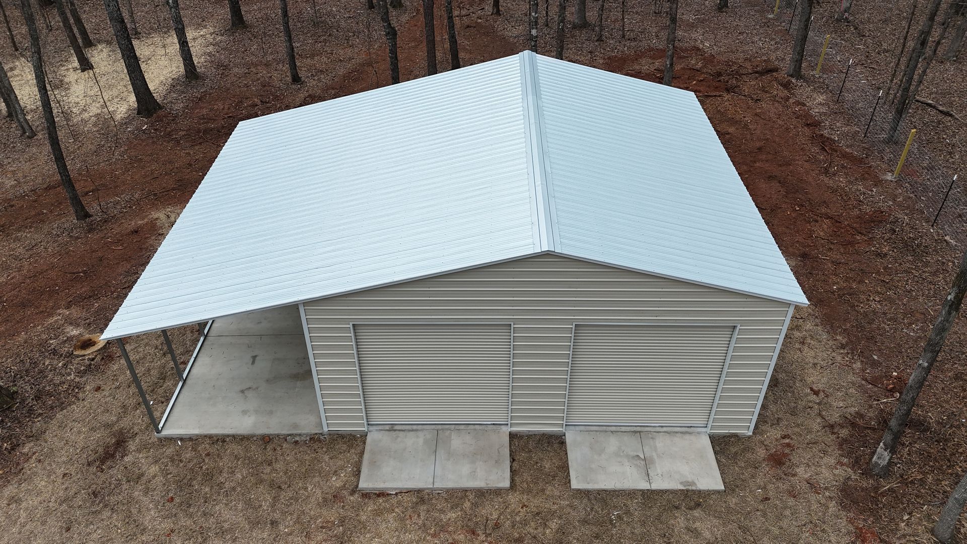 A beige shed with a white metal roof on a concrete pad, surrounded by leafless trees.