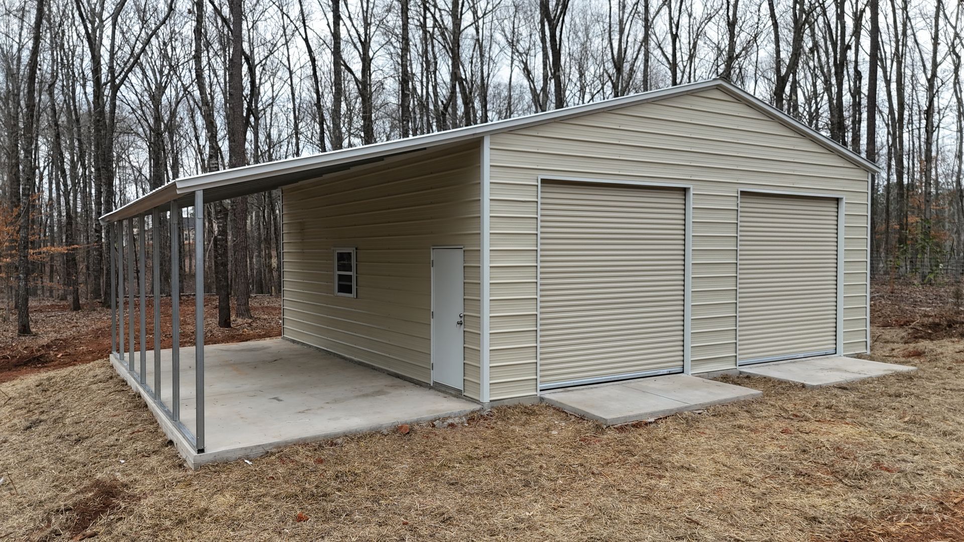 Tan metal storage shed with concrete slab and open lean-to, set in a wooded yard.