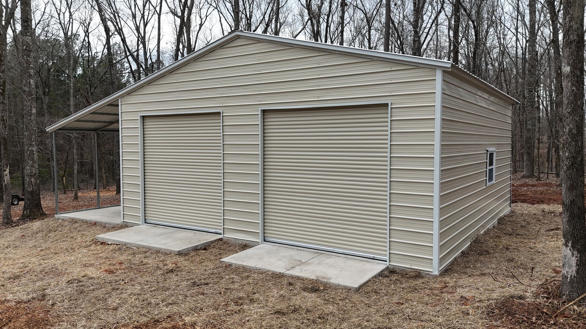 Tan metal storage building with two roll-up doors in a wooded yard