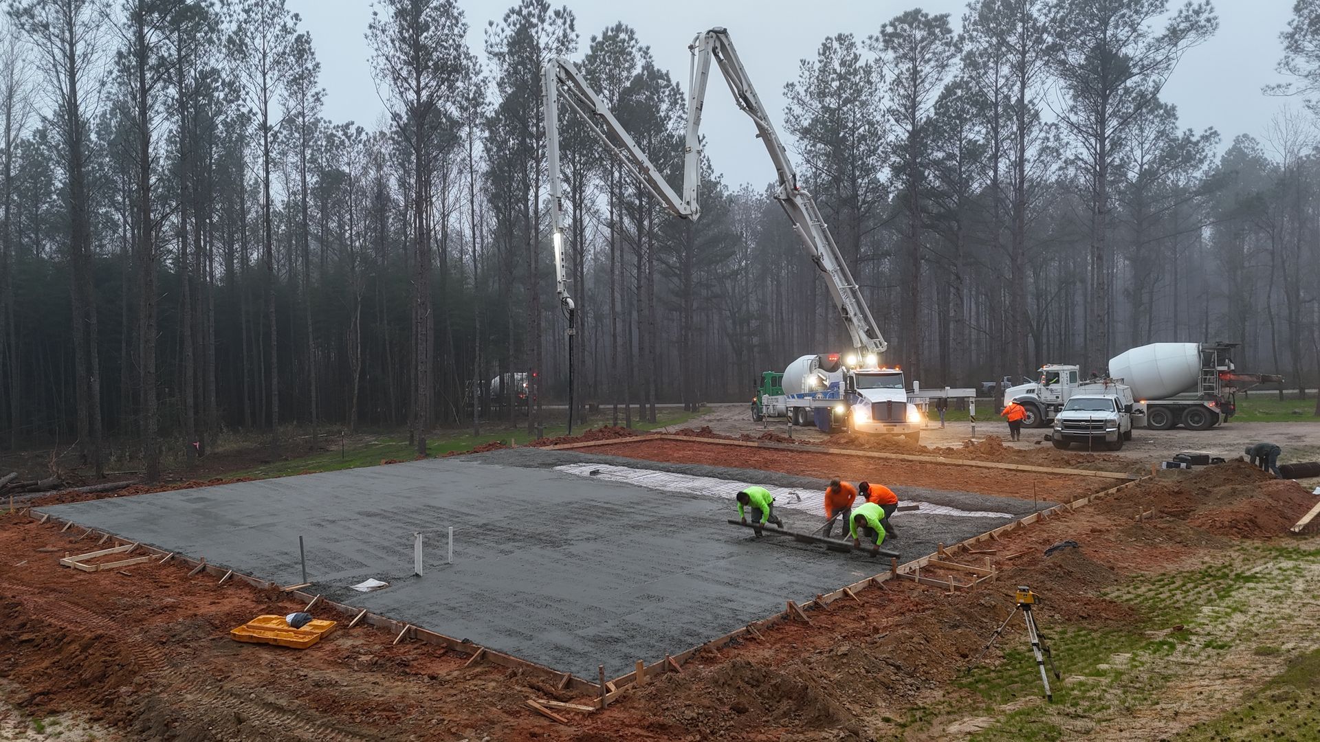 Construction crew pouring a concrete slab at a wooded worksite, with trucks and a cement mixer nearby.