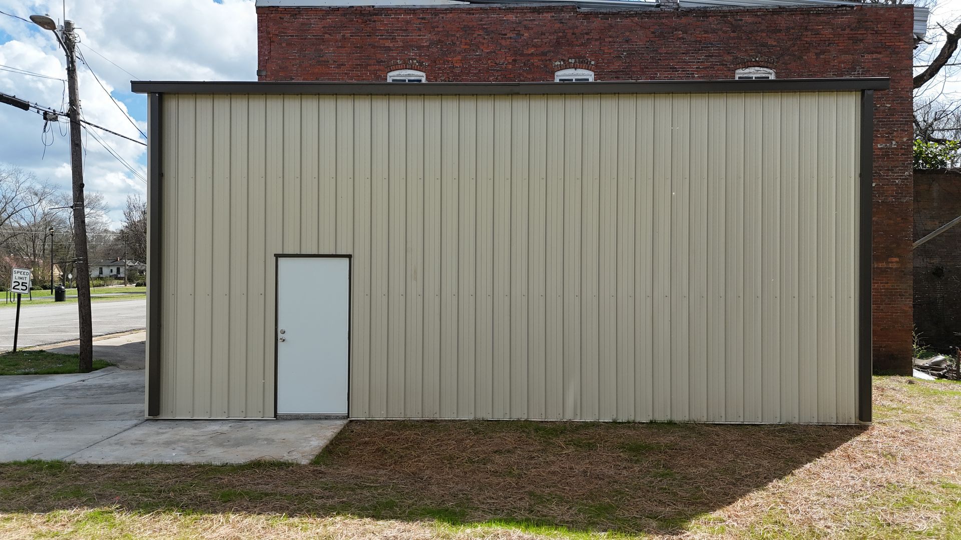 A tan metal building exterior with a single white door, positioned next to an older red brick structure.