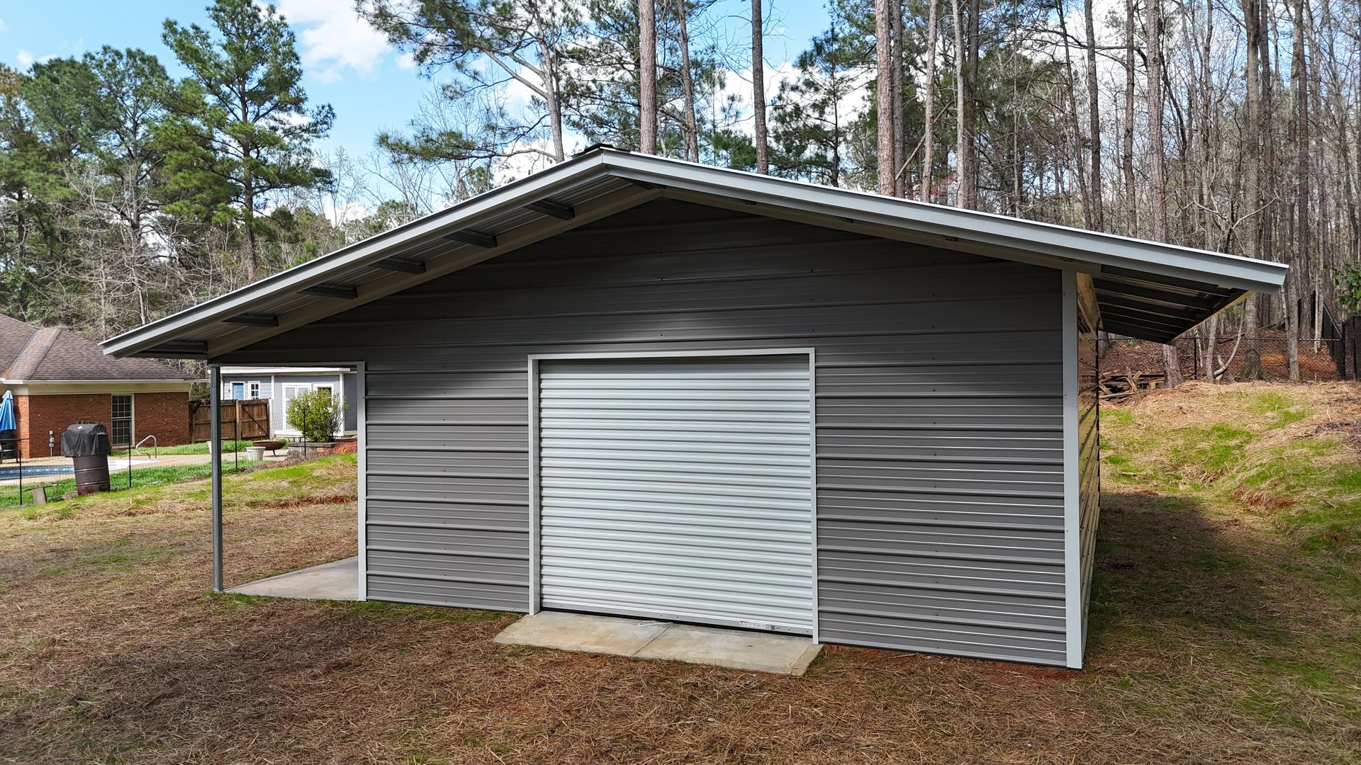 A grey metal storage building with a roll-up garage door and an attached open carport, situated in a wooded yard.