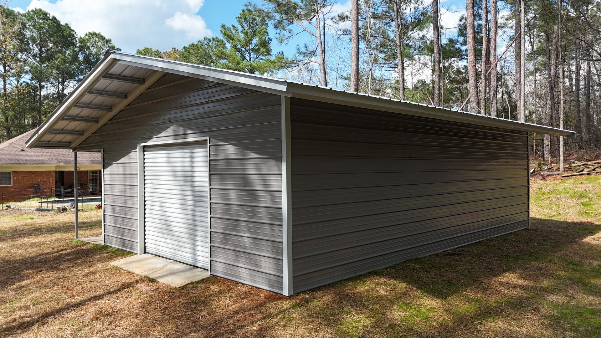 A grey metal storage building with a roll-up door stands on a grassy lot near a wooded area.