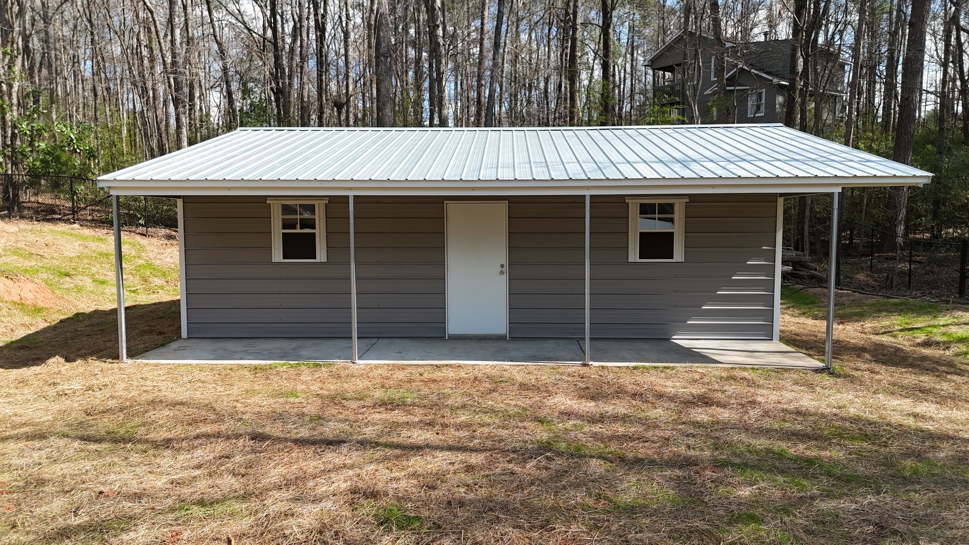 A one-story metal-sided shed with a white door, two windows, and a covered porch in a wooded, grassy outdoor setting.