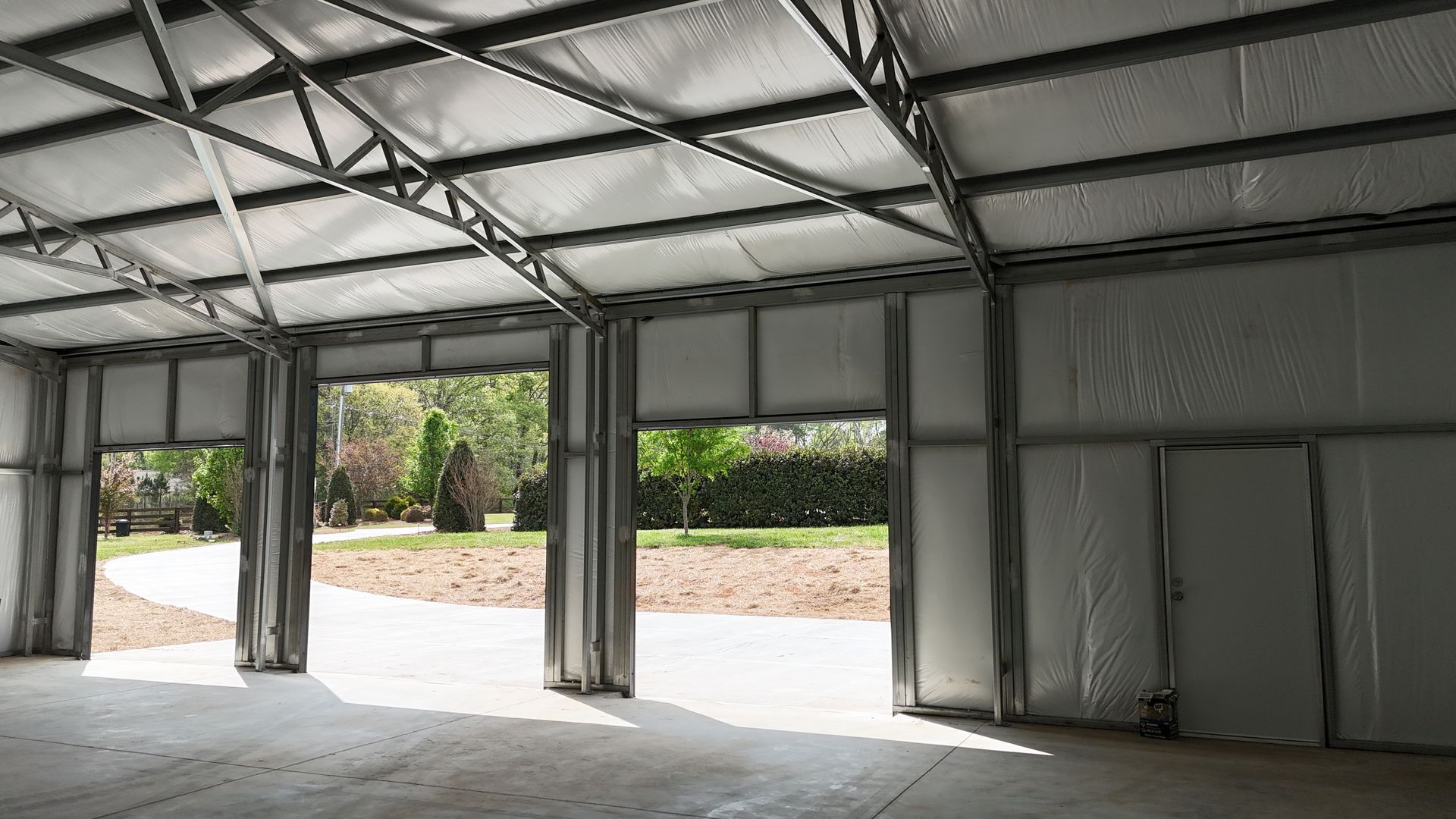 Empty metal-framed warehouse tent with open sides and concrete floor, showing grass and trees outside.