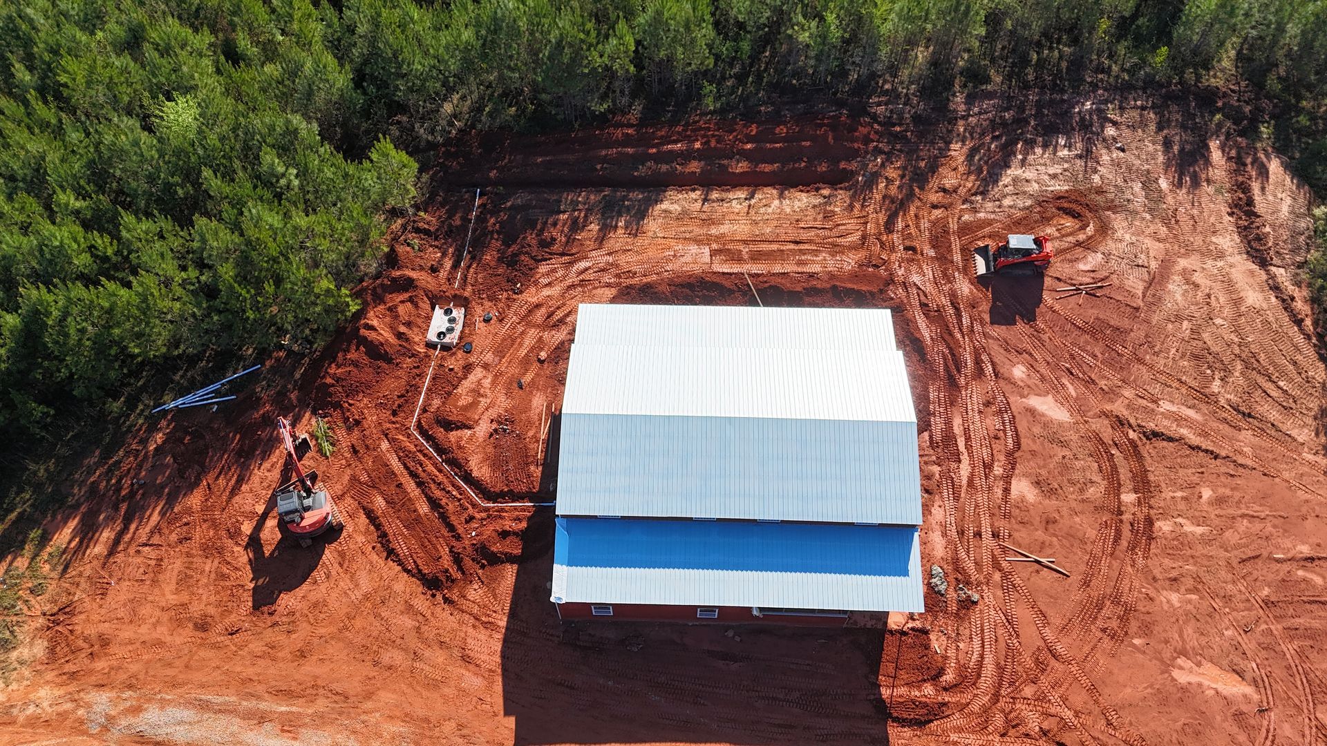 Aerial view of a small white-roofed building on red dirt beside dense green trees