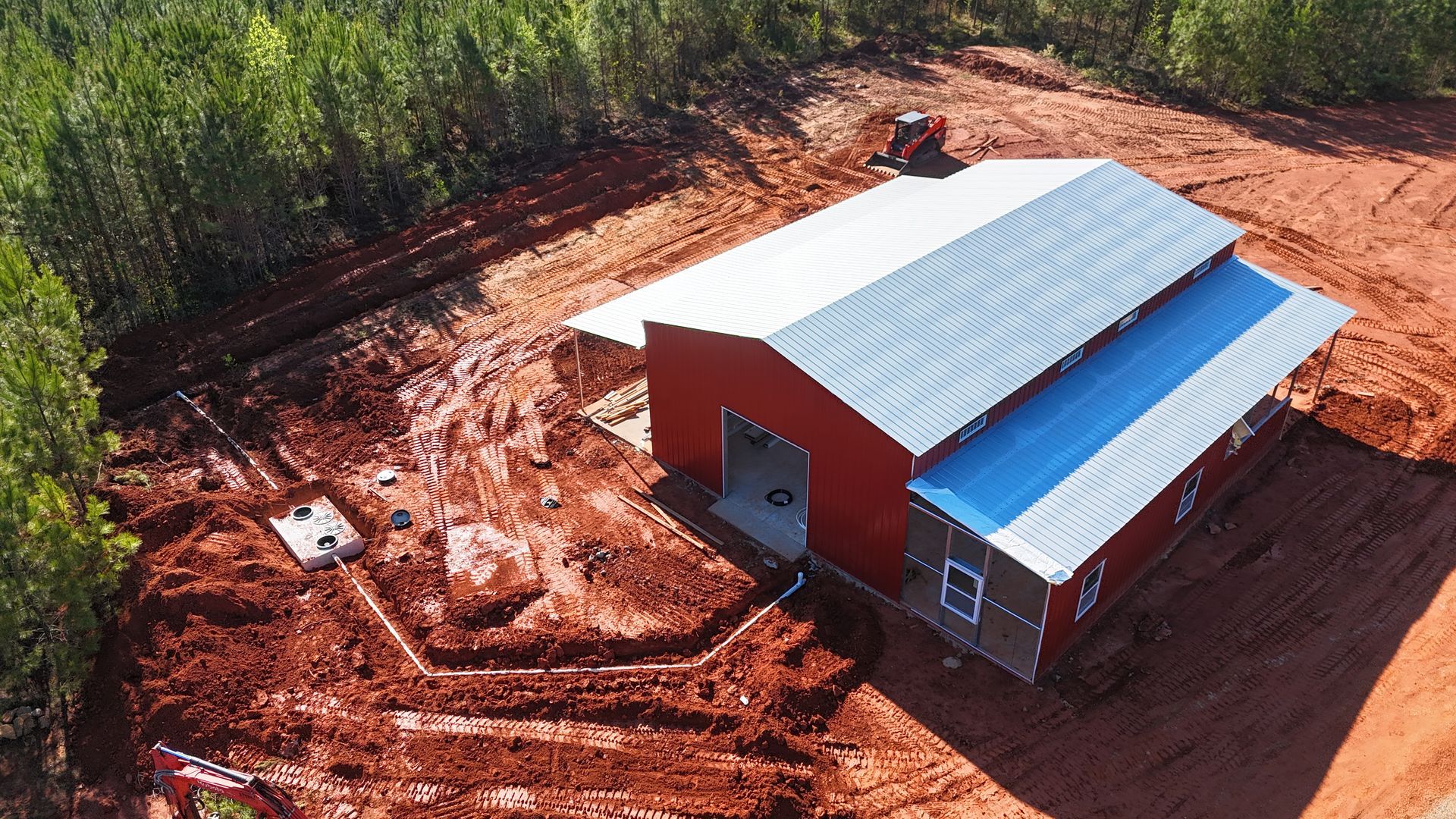 Aerial view of a red building with a white roof on a cleared dirt lot beside trees