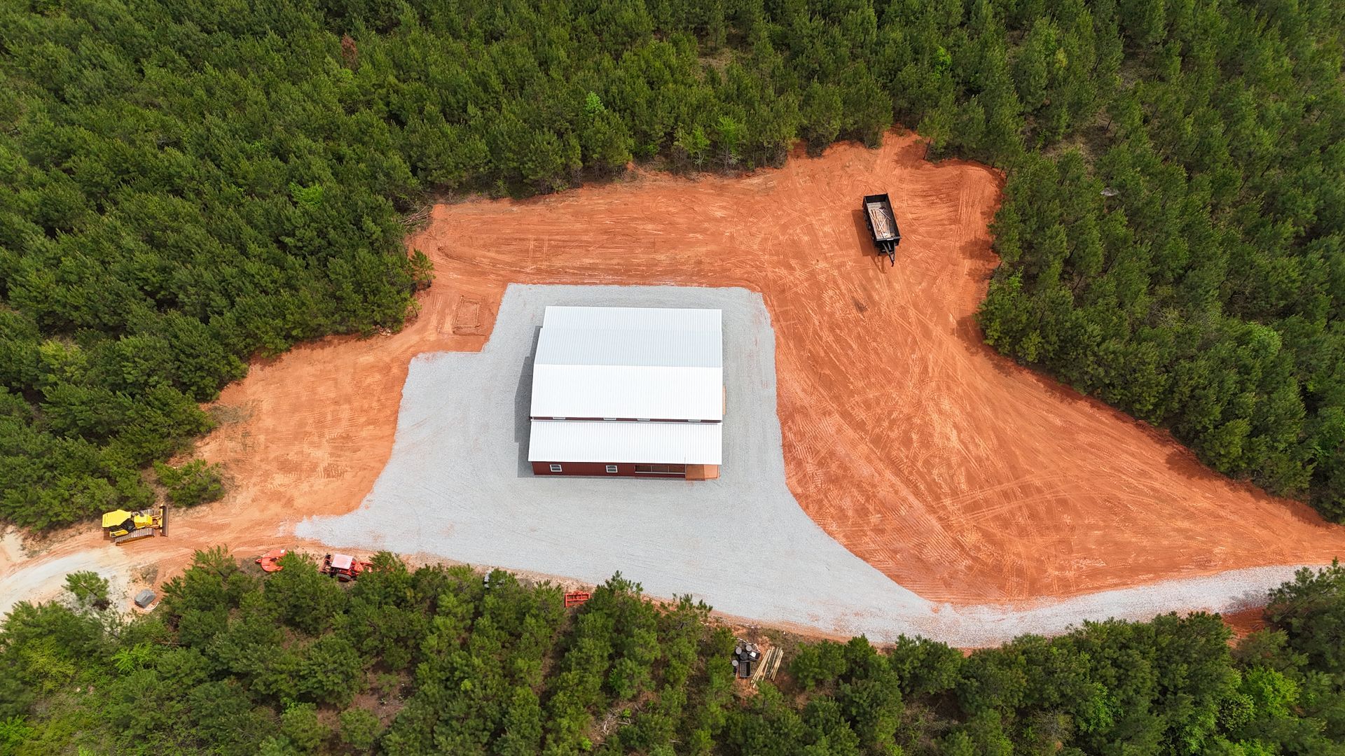 Aerial view of a white building in a cleared red dirt lot surrounded by dense green forest, with a vehicle nearby