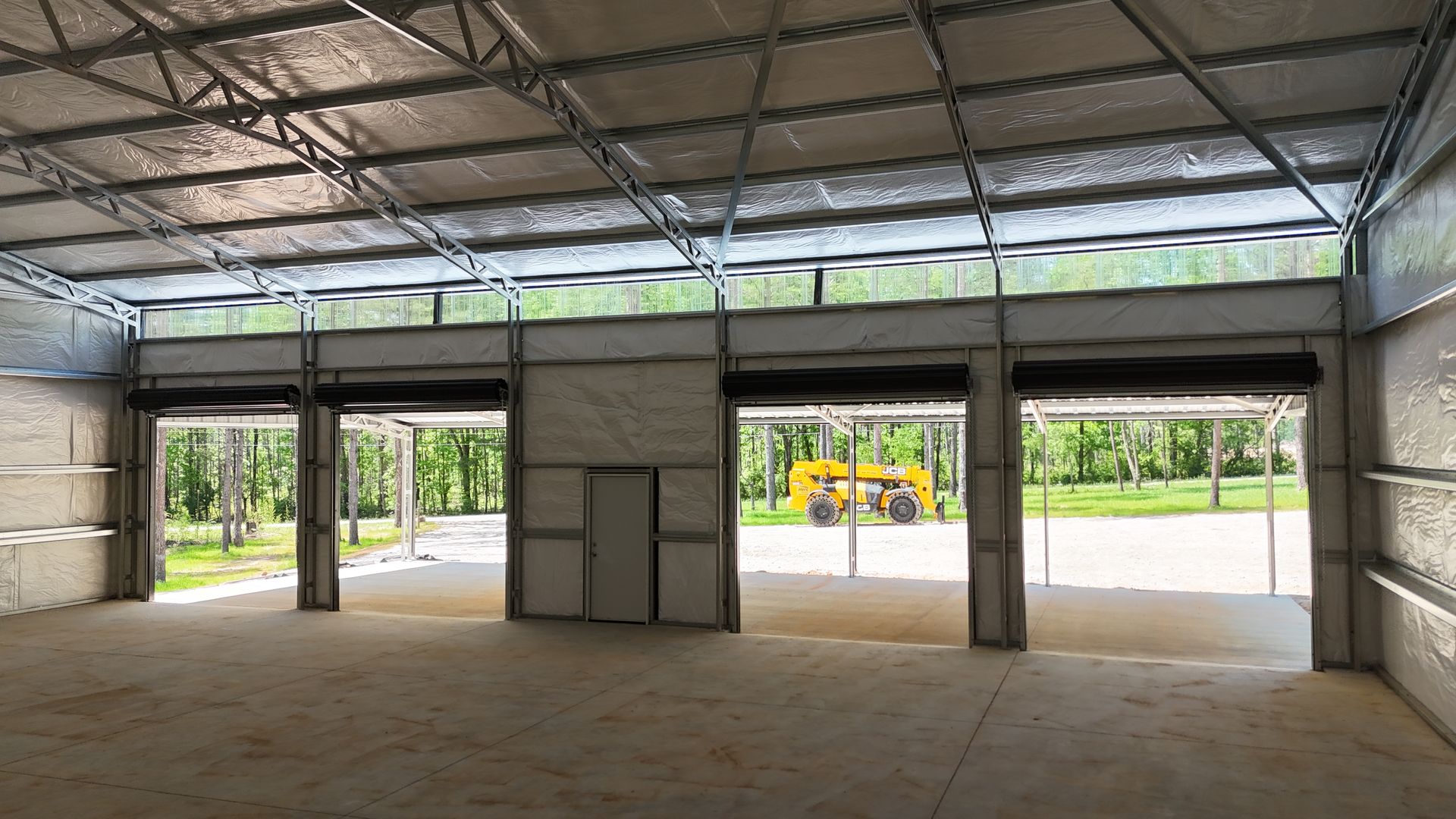 Empty open-sided warehouse interior with concrete floor and metal roof, looking out to a road and greenery.