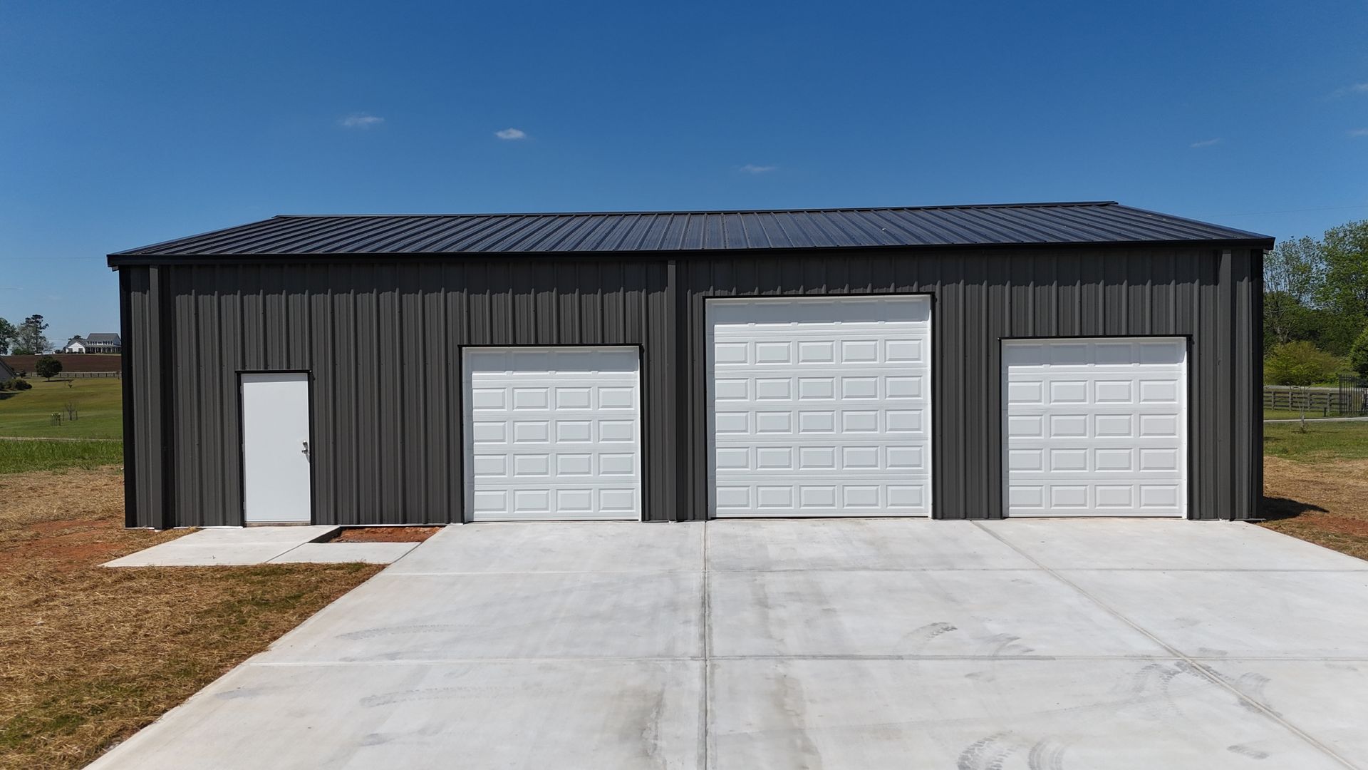 Black metal building with three garage doors and a white side entrance, viewed from a concrete driveway