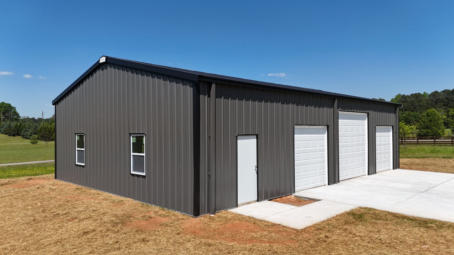 Dark gray metal warehouse building with white garage doors and a concrete pad in a rural setting