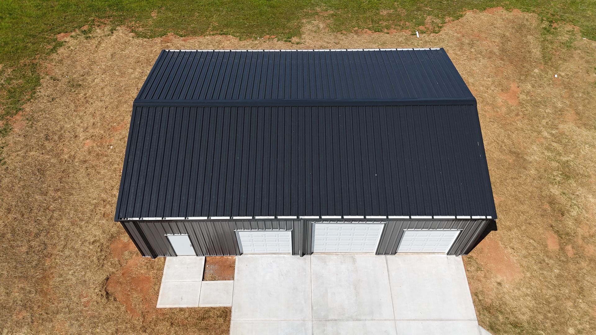 Aerial view of a modern black-roofed house with a white concrete driveway and dry grass yard.