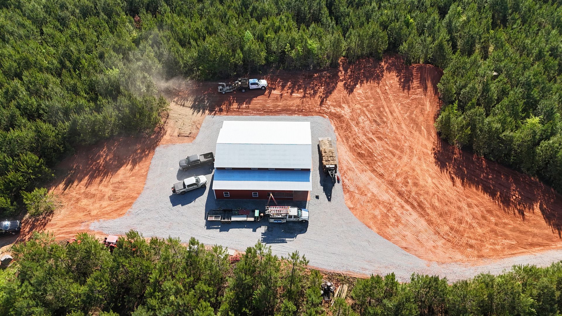 Aerial view of a white-roofed building and parked cars on a gravel lot beside red dirt clearing and trees