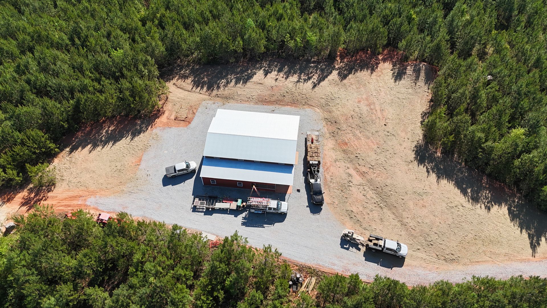 Aerial view of a white-roofed building and two vehicles beside a gravel lot in a wooded area