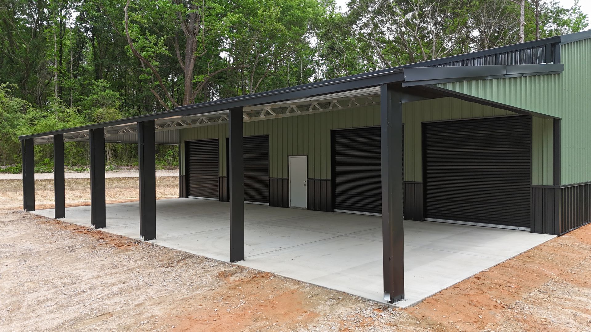 Covered concrete driveway beside a green metal garage with dark support posts and wooded background