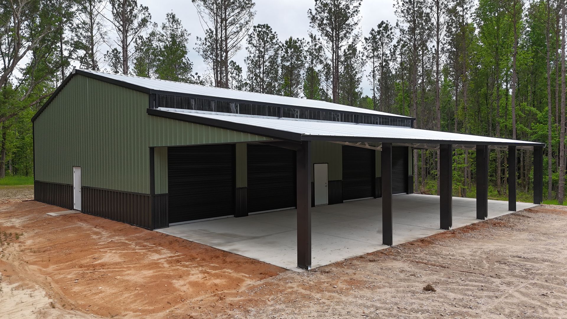 Green metal barn with covered open bays on a concrete slab in a wooded rural setting