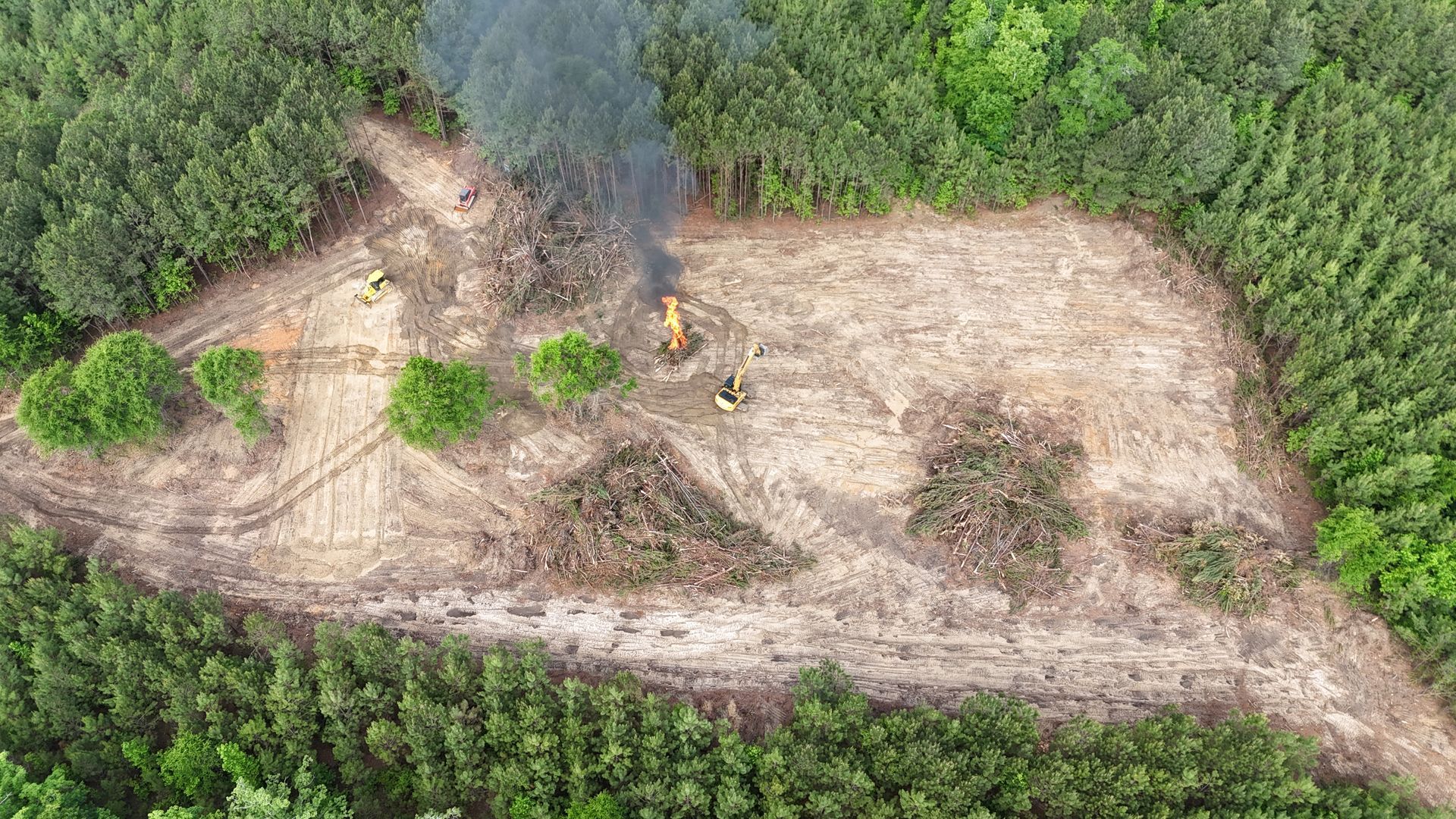 Aerial view of a forest clearing with a small fire and smoke, surrounded by dense green trees.