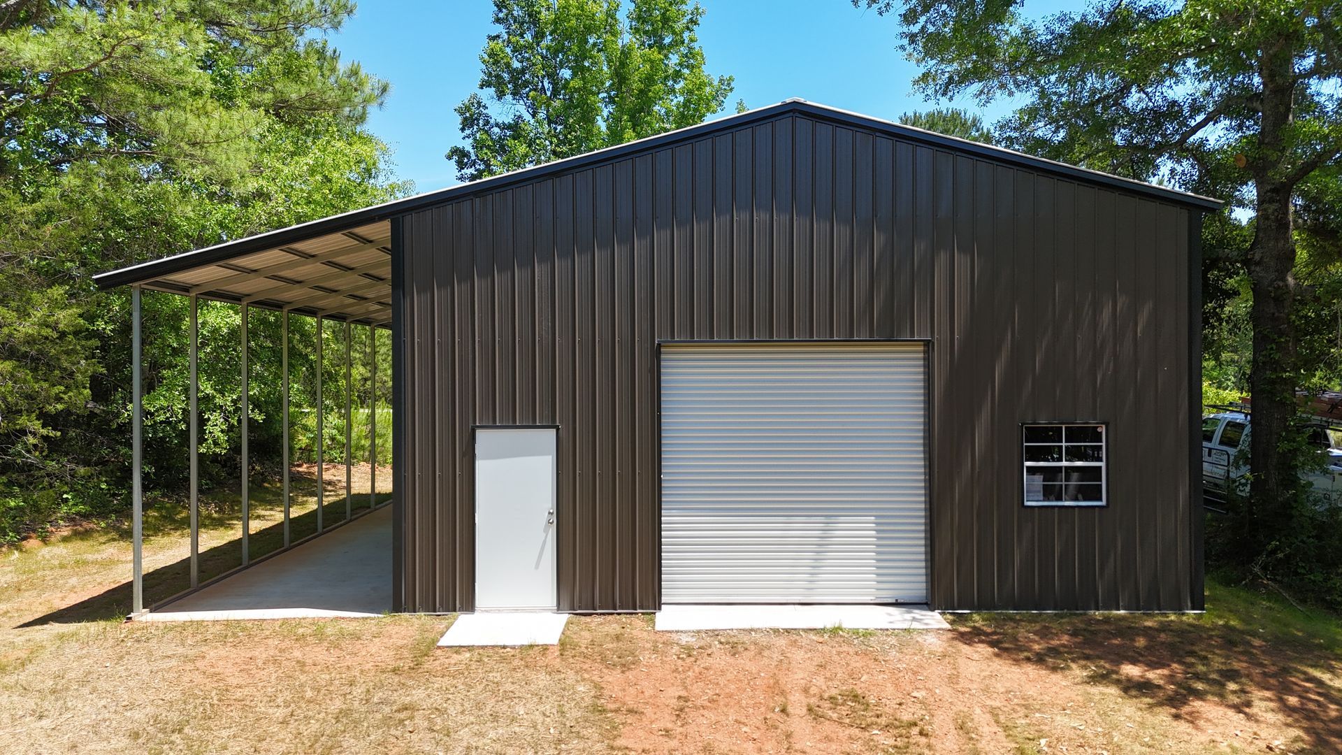 Dark metal shed with a white garage door and side door, beside a covered open bay in a wooded yard.