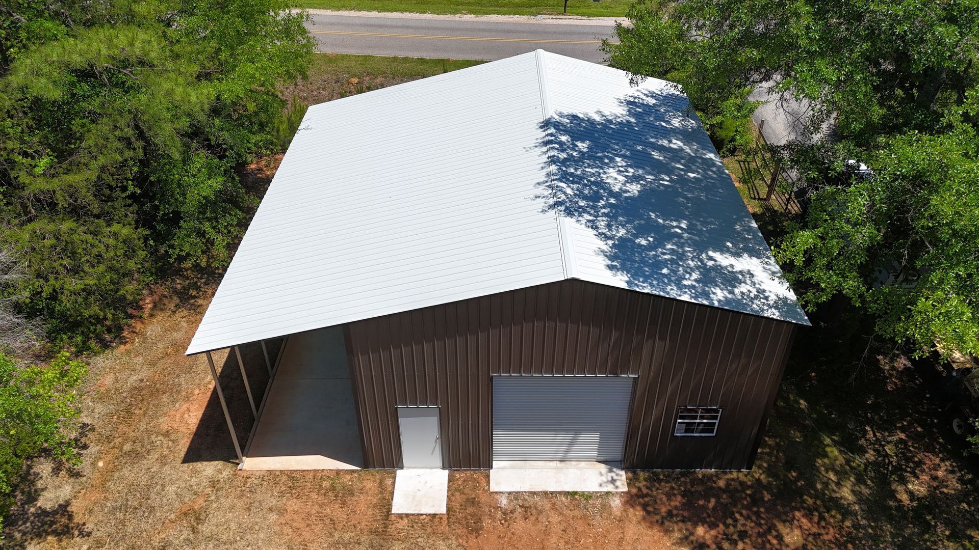 Aerial view of a small brown metal building with a white roof beside a road and trees.