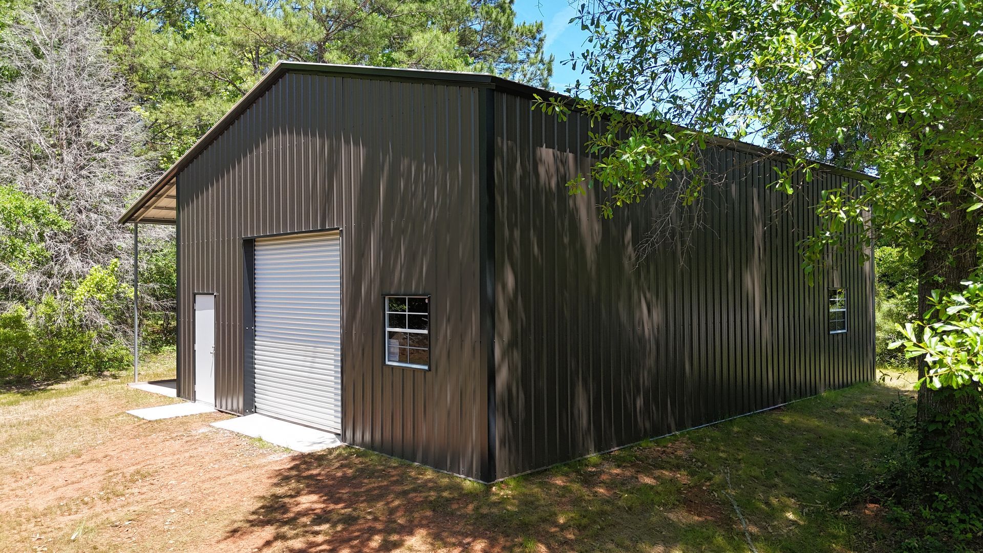 Dark metal shed with a white roll-up door and small window, surrounded by trees in a wooded area