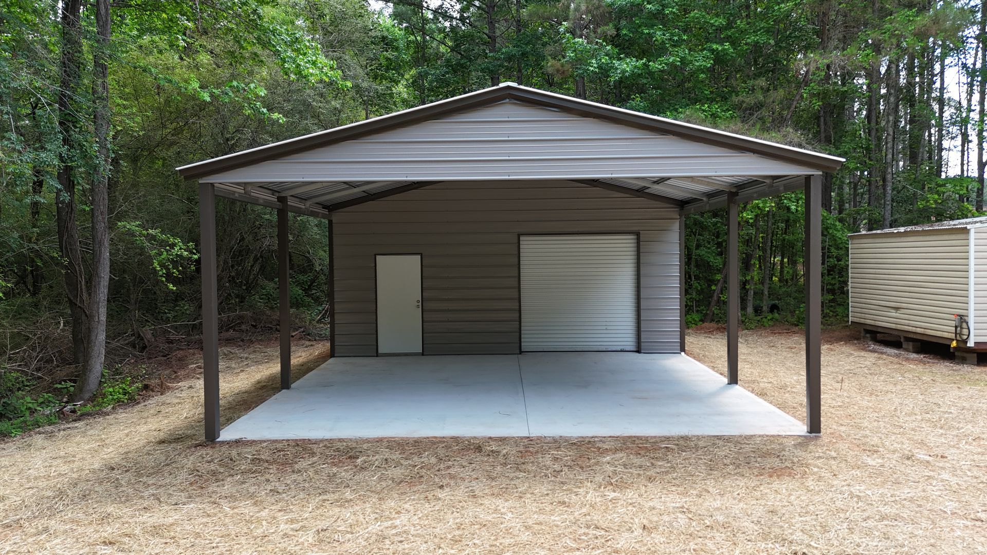 Open-front metal carport with two storage doors, set on gravel beside a wooded area.