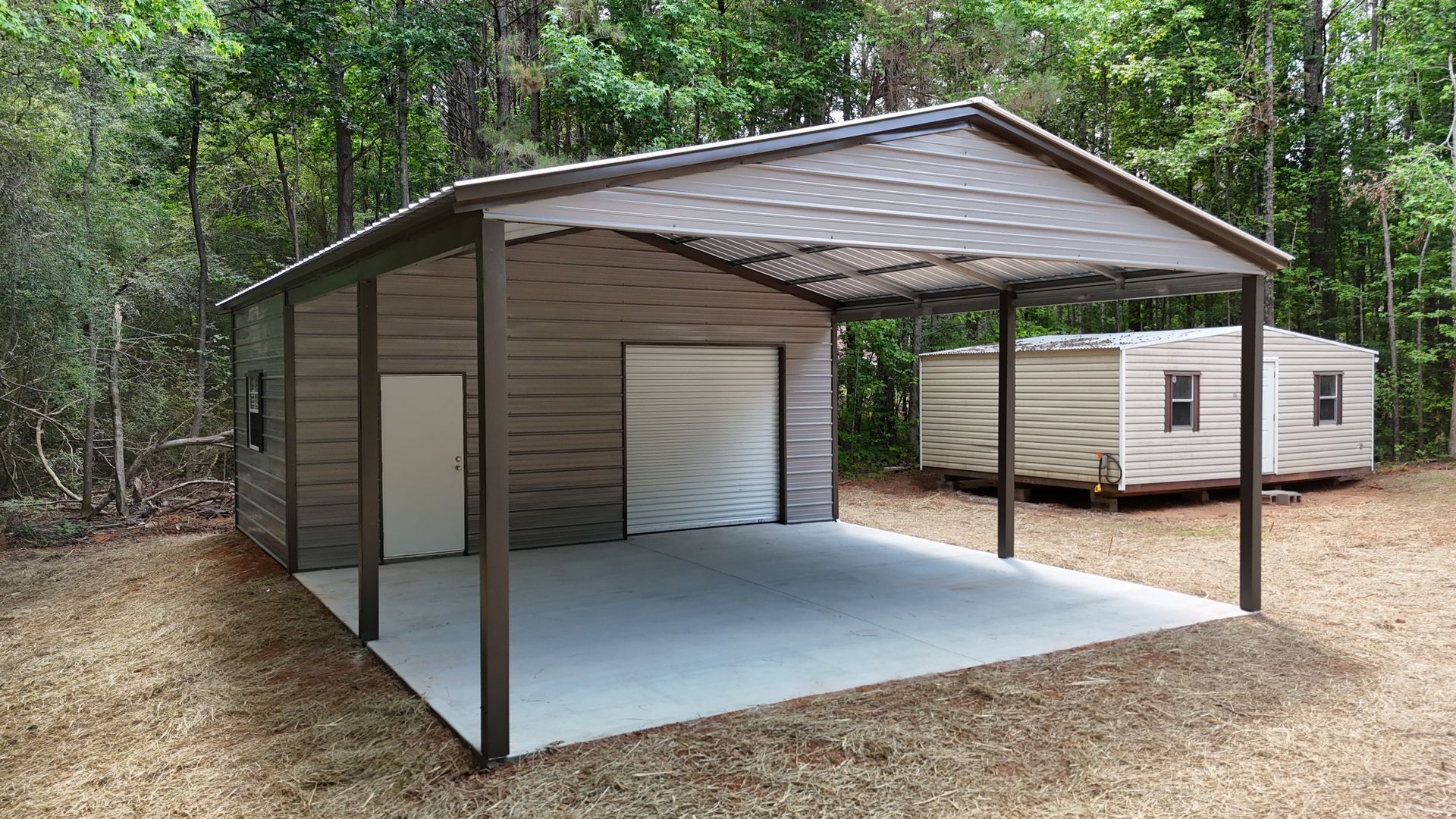 Open-sided metal carport with a concrete floor in a wooded yard, beside a white storage shed.