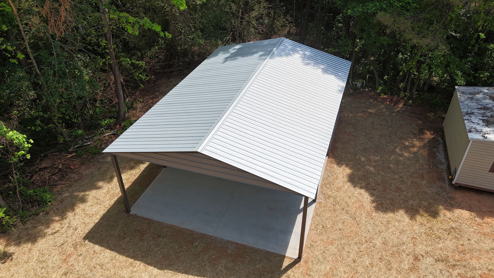 White metal-roofed shelter on bare ground in a wooded outdoor area