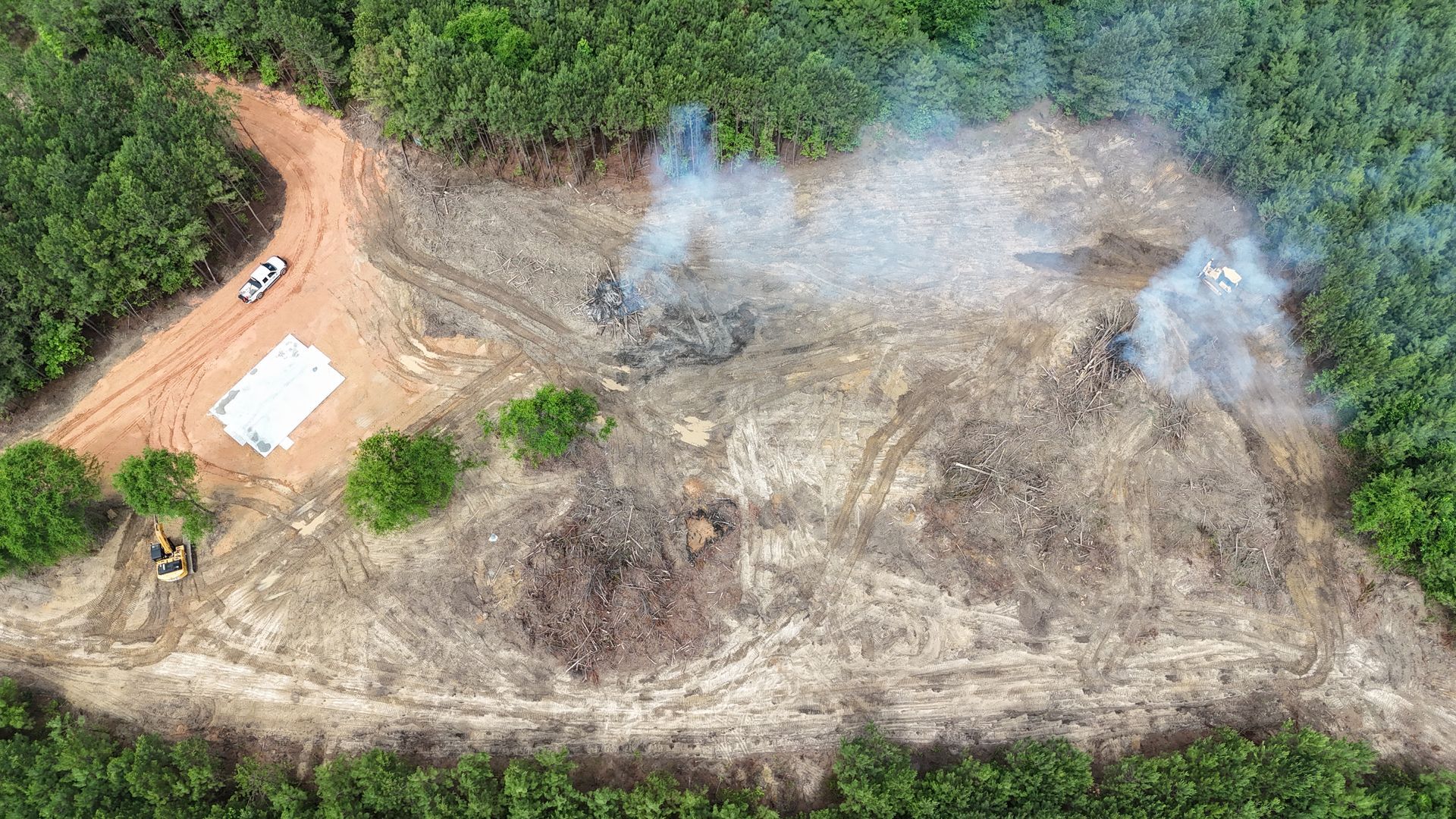 Aerial view of a cleared dirt lot with smoke rising, surrounded by dense green trees.