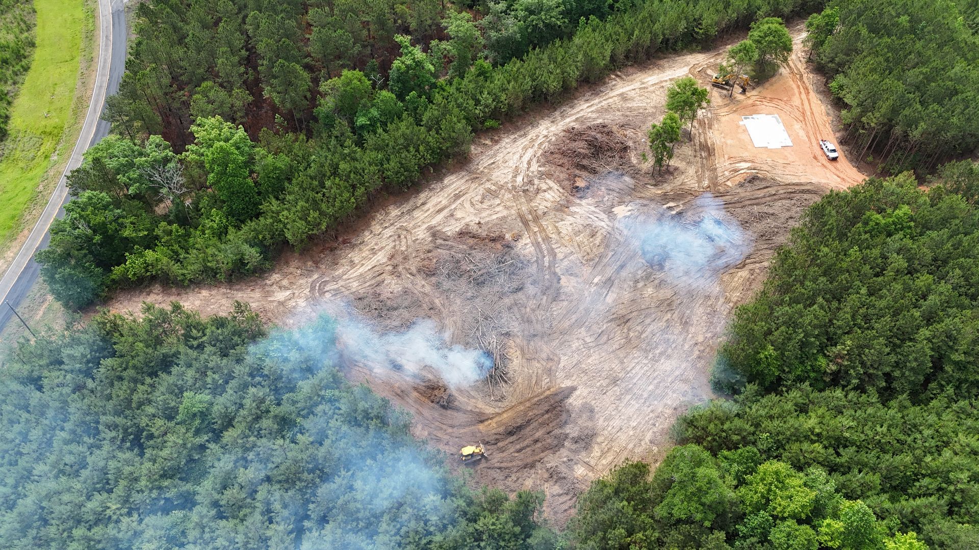Aerial view of a burned clearing bordered by dense green forest, with smoke rising from charred ground.