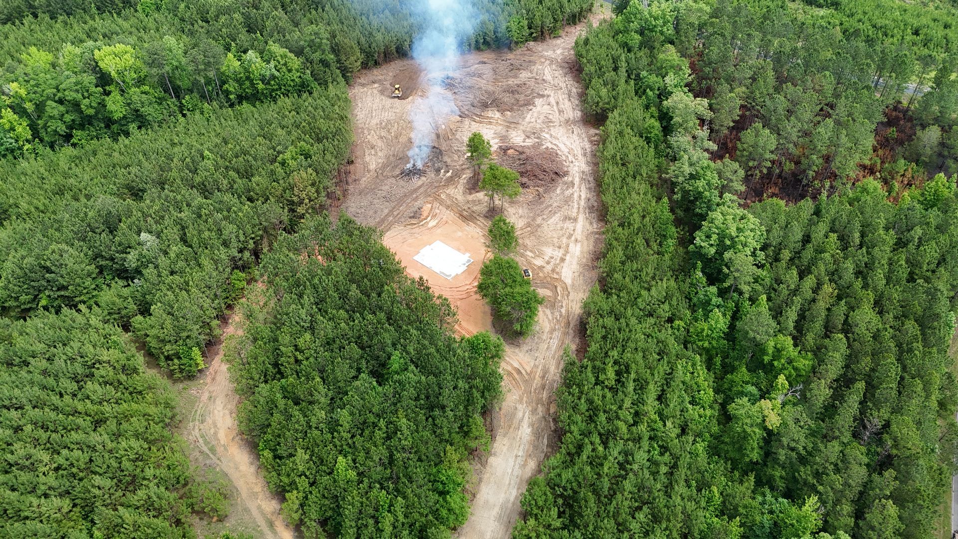 Aerial view of a dirt path through dense green forest with a small campfire or smoke plume near the center
