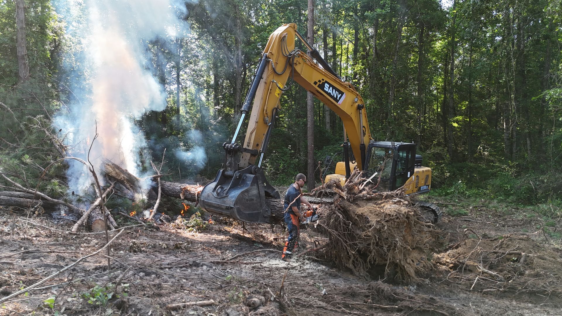 Excavator clearing a smoking debris pile in a wooded area, with workers nearby.